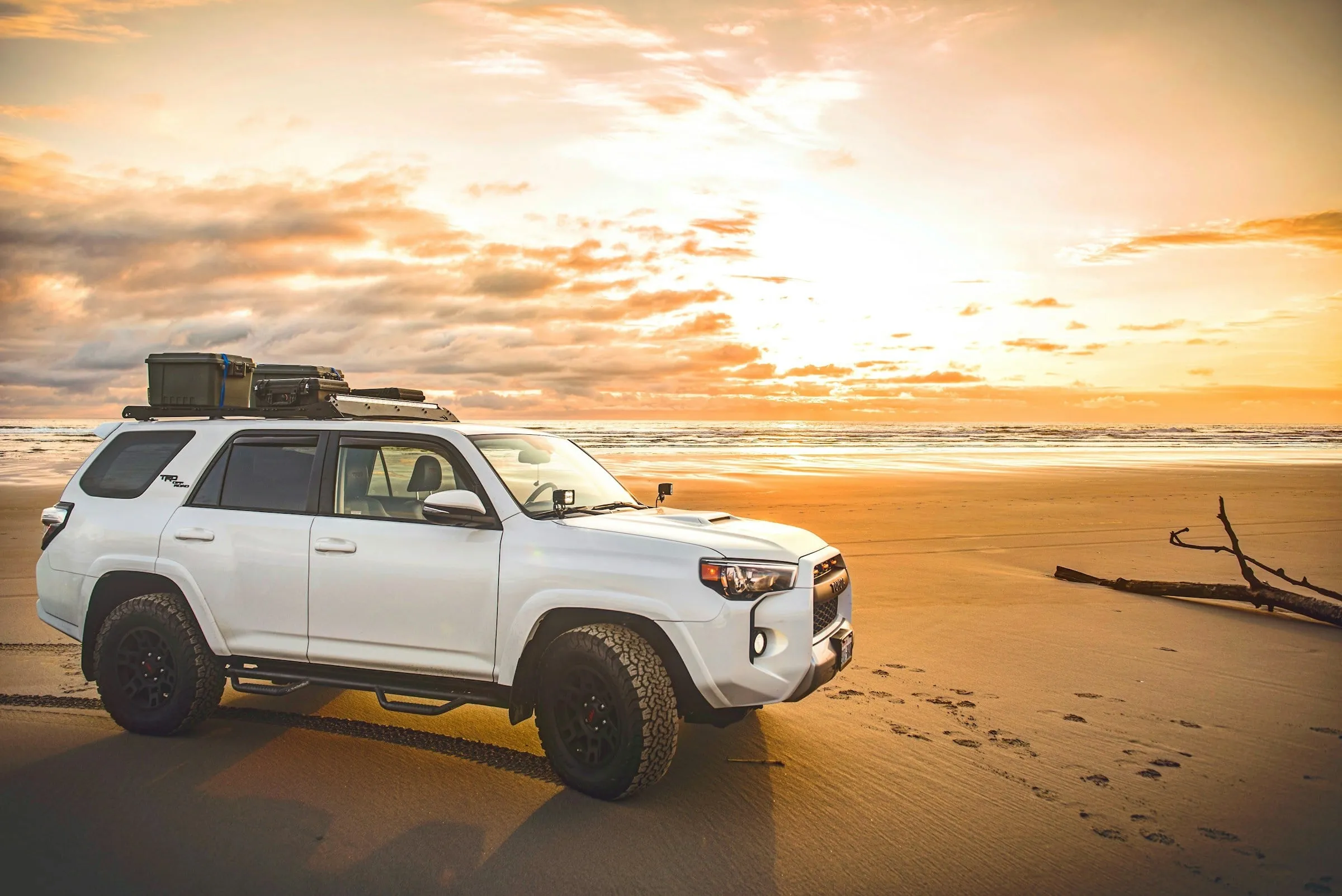 A white Toyota 4Runner TRD with a roof rack and cargo box parked on a beach at sunset