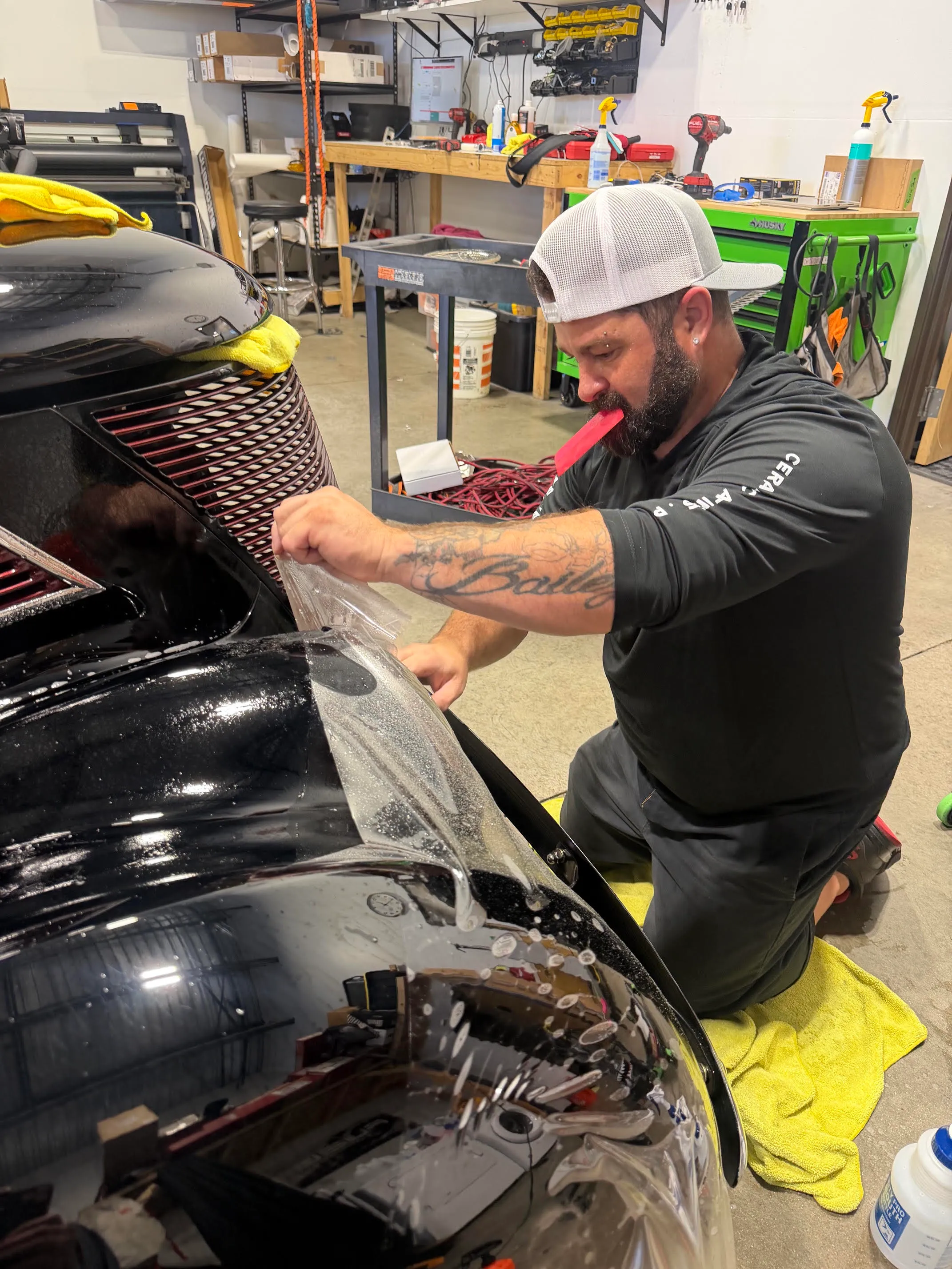 Chris Culpepper kneeling beside a black car in the shop, working a sheet of paint protection film onto the front bumper