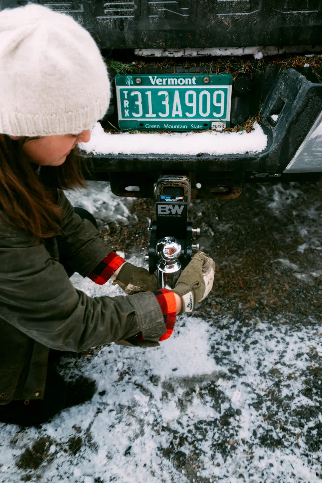 A B&W Tow & Stow ball mount in a truck receiver, its shank stamped with the B&W Trailer Hitches name, being adjusted by hand over snowy ground