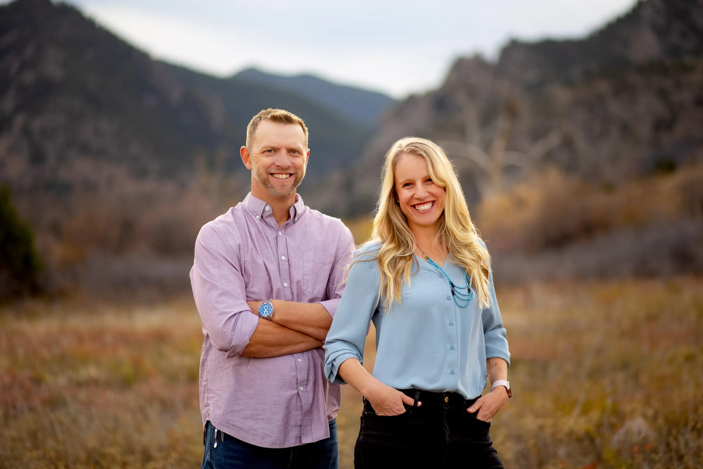 Eric and Kelsey Carlson standing together in a Colorado foothills meadow, rock outcrops behind them