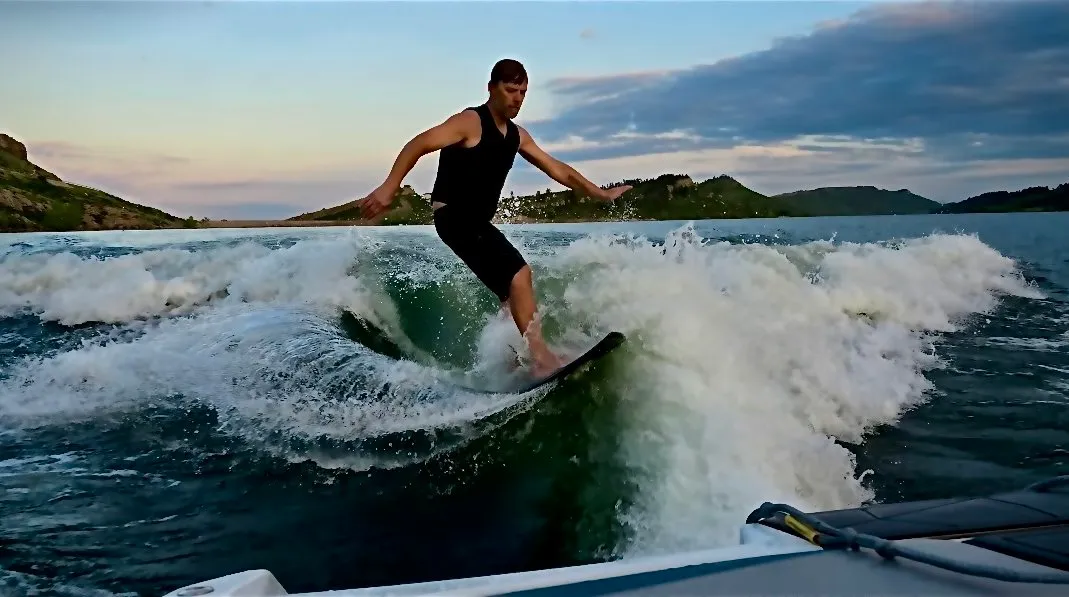 Eric Carlson wakesurfing behind a boat on a Colorado lake at dusk, hills along the far shore