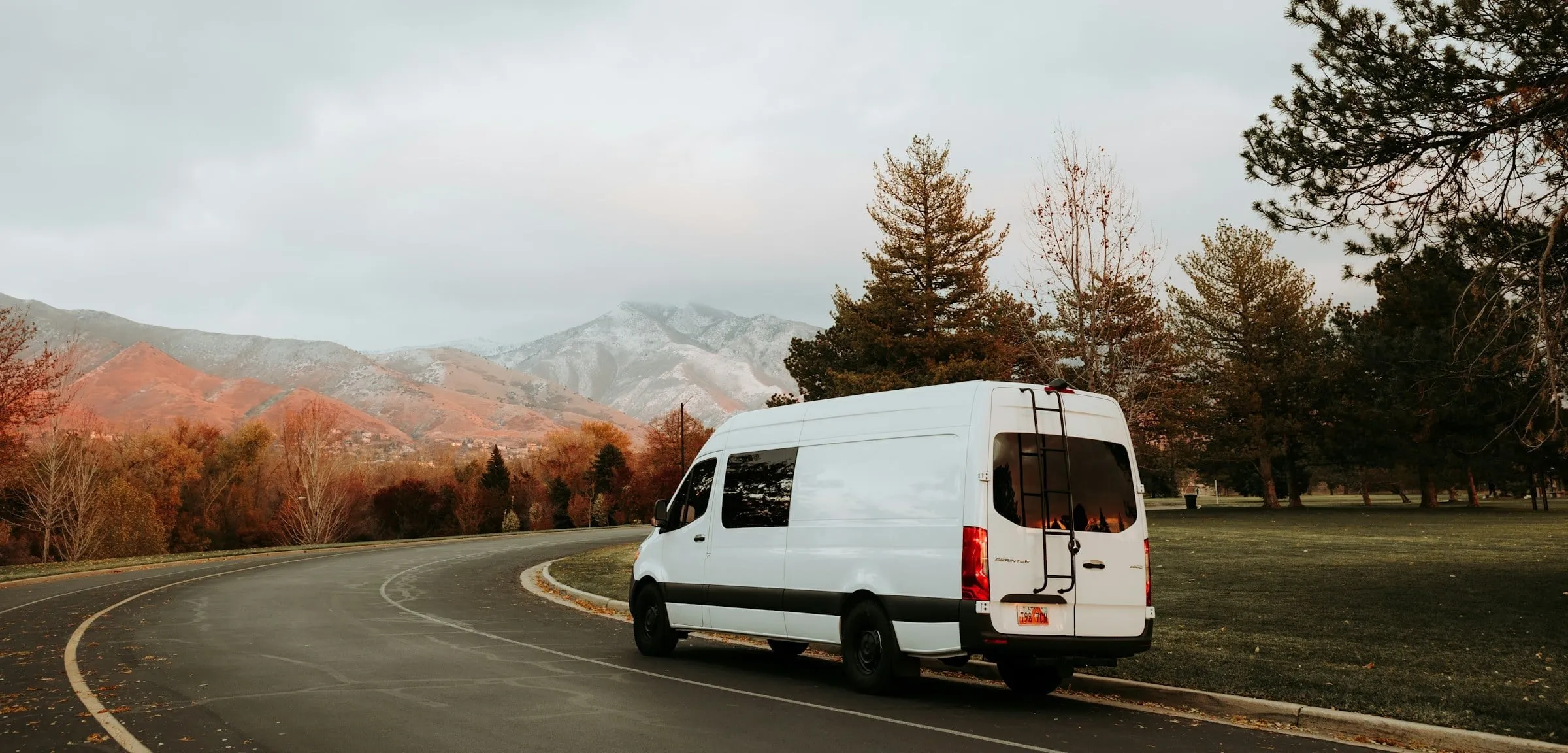 A white high-roof cargo van driving a two-lane road through autumn foothills