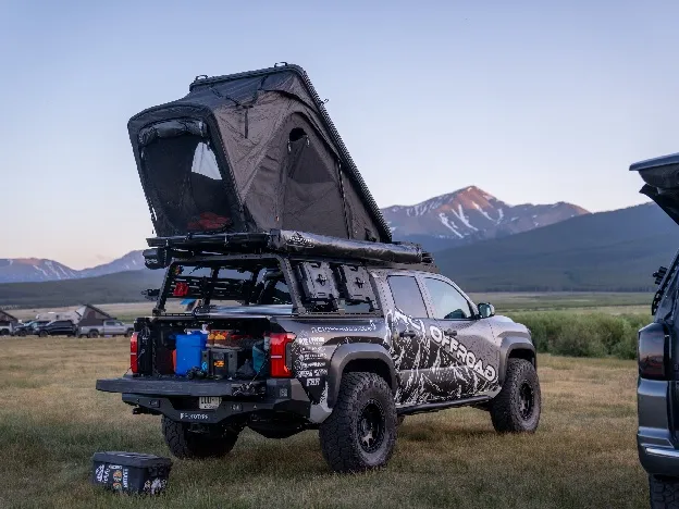 A grey Toyota Tacoma in a high mountain meadow with a hard-shell roof-top tent opened on its bed rack and drawers pulled out over the tailgate