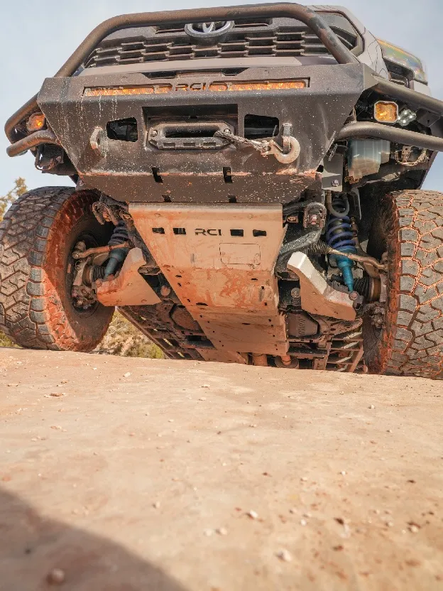 The underside of a Toyota Tacoma on red rock, showing an RCI front bumper with winch and light bar, aluminum skid plates, and mud-caked all-terrain tires