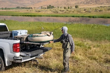 A fly fisherman lifting a float tube from the open tailgate of a white pickup beside a river, with a cooler and gear sorted in the bed