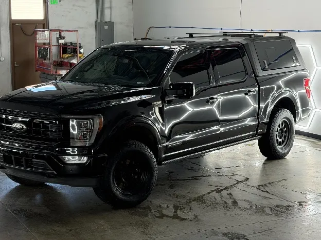 A gloss black Ford F-150 with a color-matched truck cap, roof rack, and black wheels parked under hexagon detailing lights