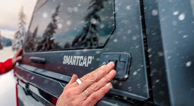 A hand pressing the key lock on the side hatch of a black truck cap while snow falls