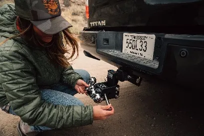 A woman in a trucker cap kneeling on a dirt road, tightening an adjustable drop hitch in the receiver of a black Nissan Titan