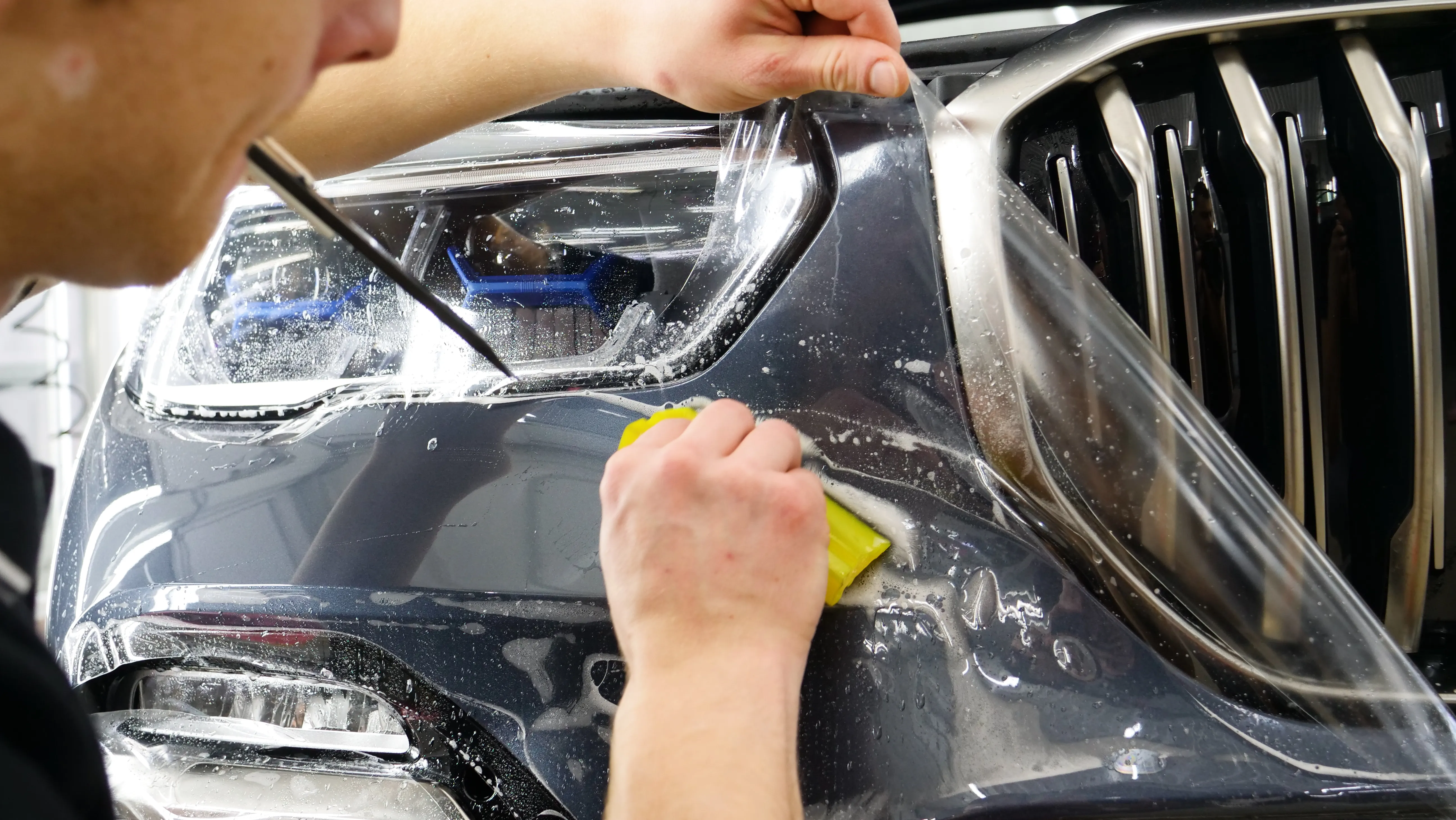 An installer squeegeeing paint protection film over the headlight and front fender of a dark grey SUV during a wet install