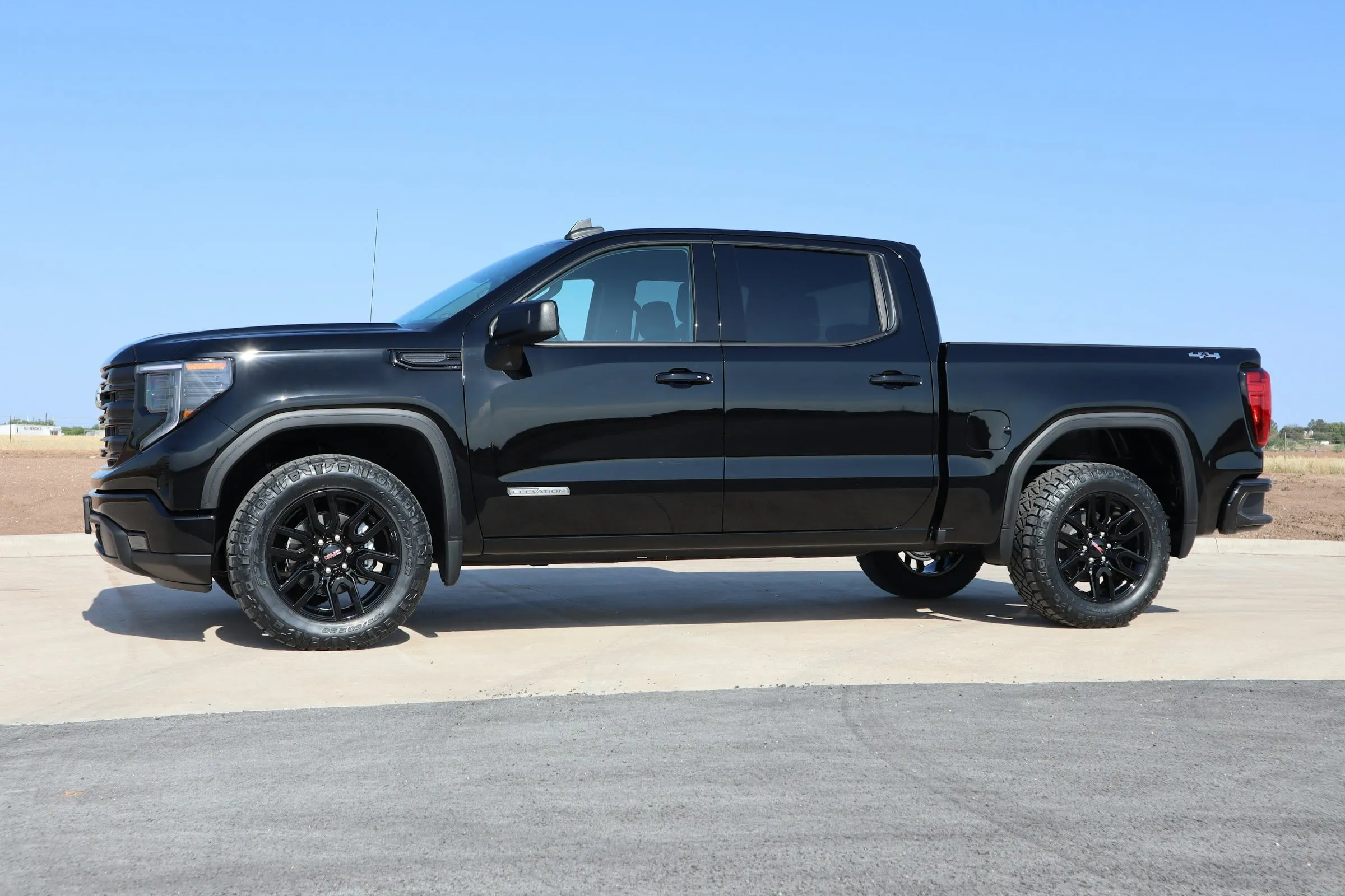 A black GMC Sierra 1500 crew cab on all-terrain tires, side view on a concrete pad under a clear sky