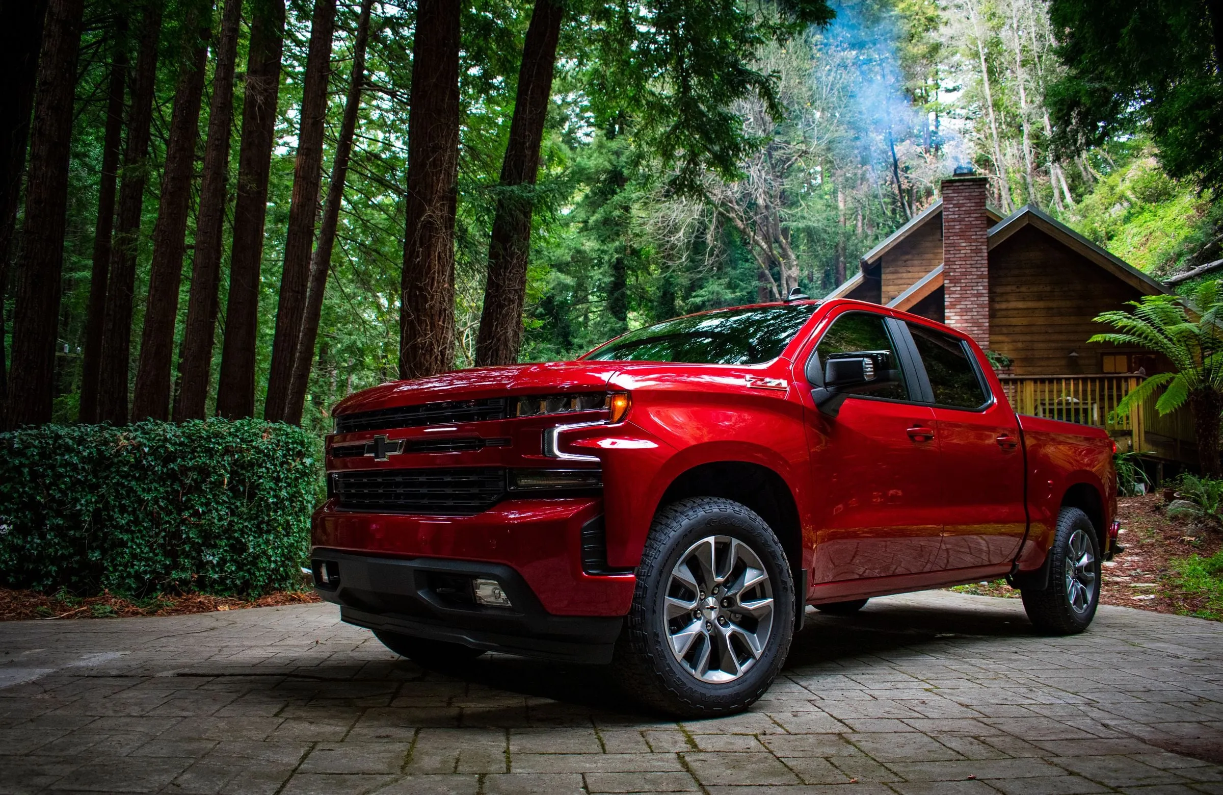 A red Chevrolet Silverado 1500 RST crew cab parked on a paved driveway in front of a cabin among tall pines