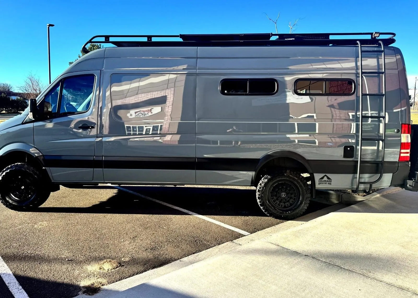 A grey lifted Mercedes Sprinter 4x4 camper van with a roof rack and rear ladder, side view in the lot after a ceramic coating