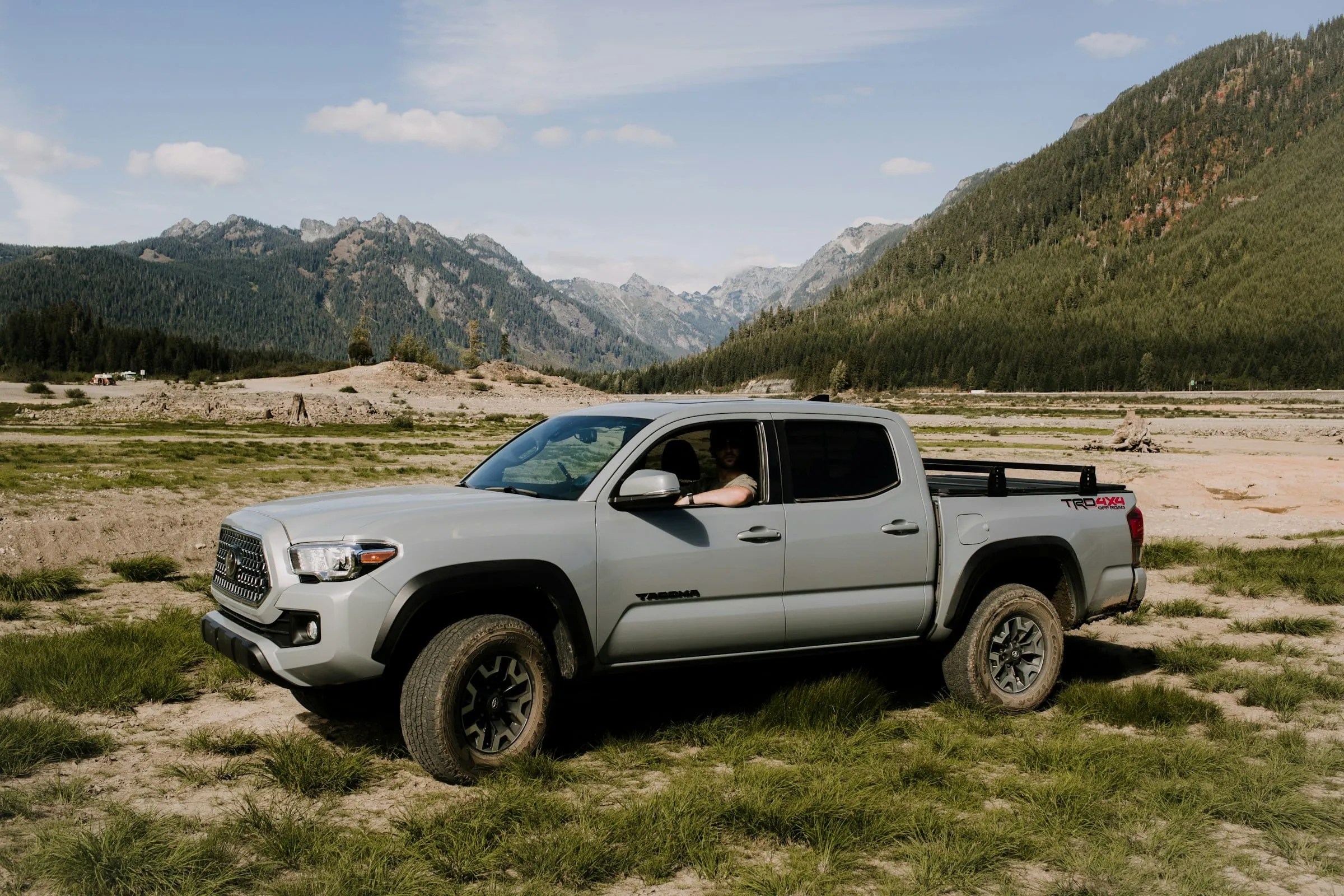 A grey Toyota Tacoma TRD Off-Road parked on an open gravel flat with forested mountains behind it