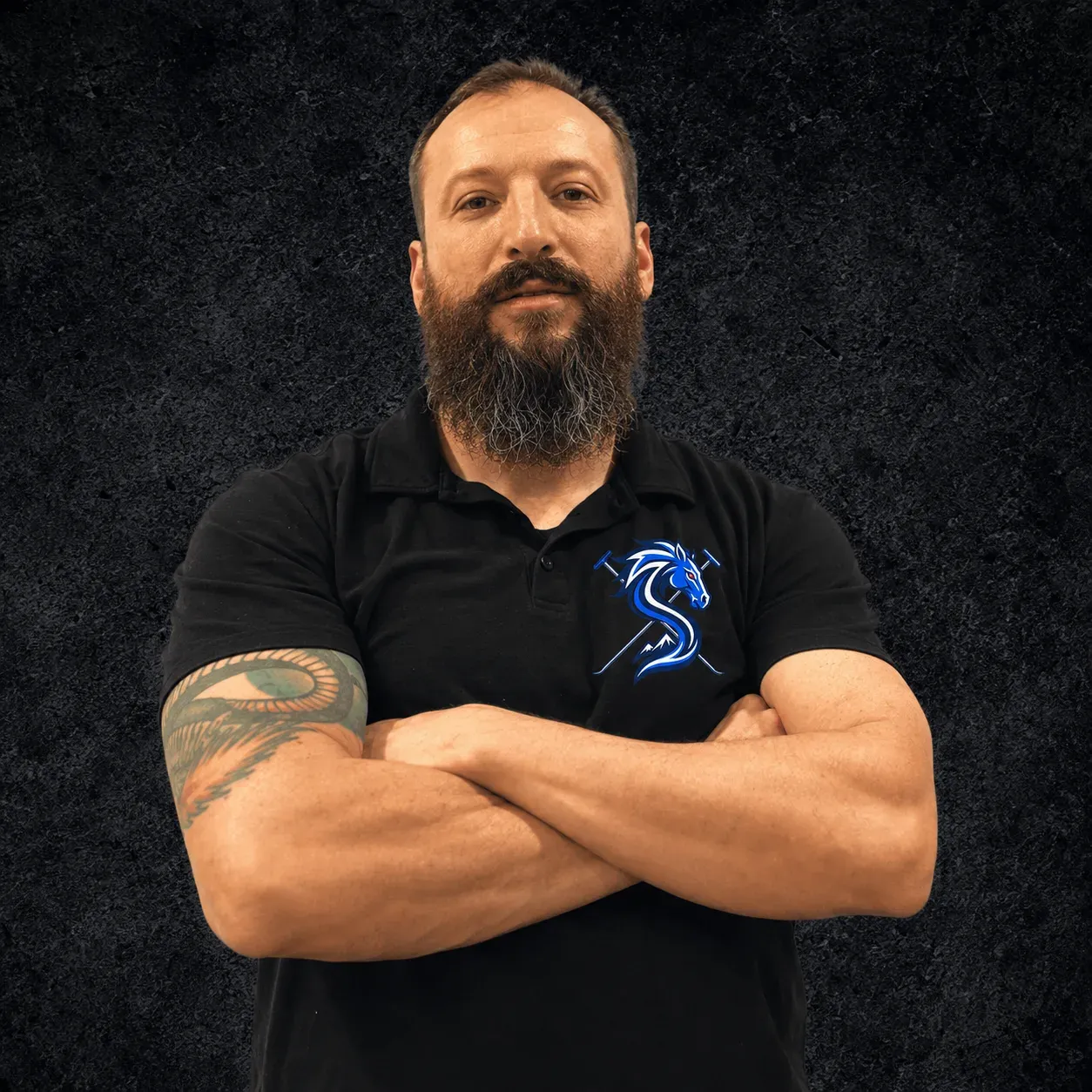 Travis Stearns with his arms crossed in a black polo shirt, photographed against a dark textured backdrop