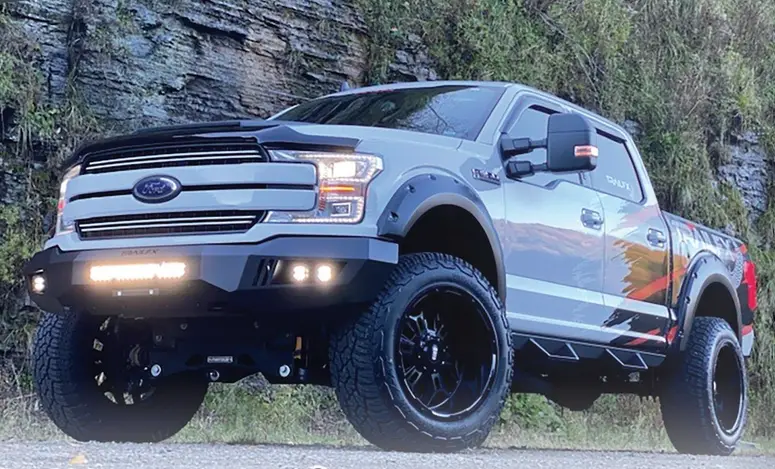 A lifted grey Ford F-150 with a steel front bumper, light bar, fender flares and black off-road wheels, parked on gravel below a rock face