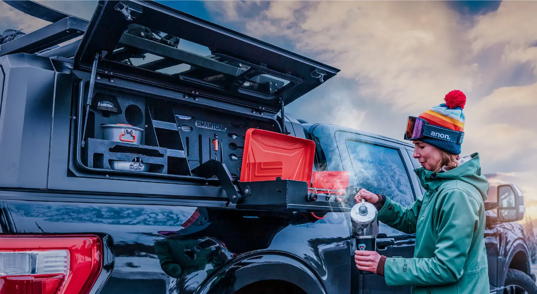 A black pickup with a truck cap and its side hatch open, a fold-out camp table and orange cooler mounted inside, while a woman in a beanie pours coffee at sunset