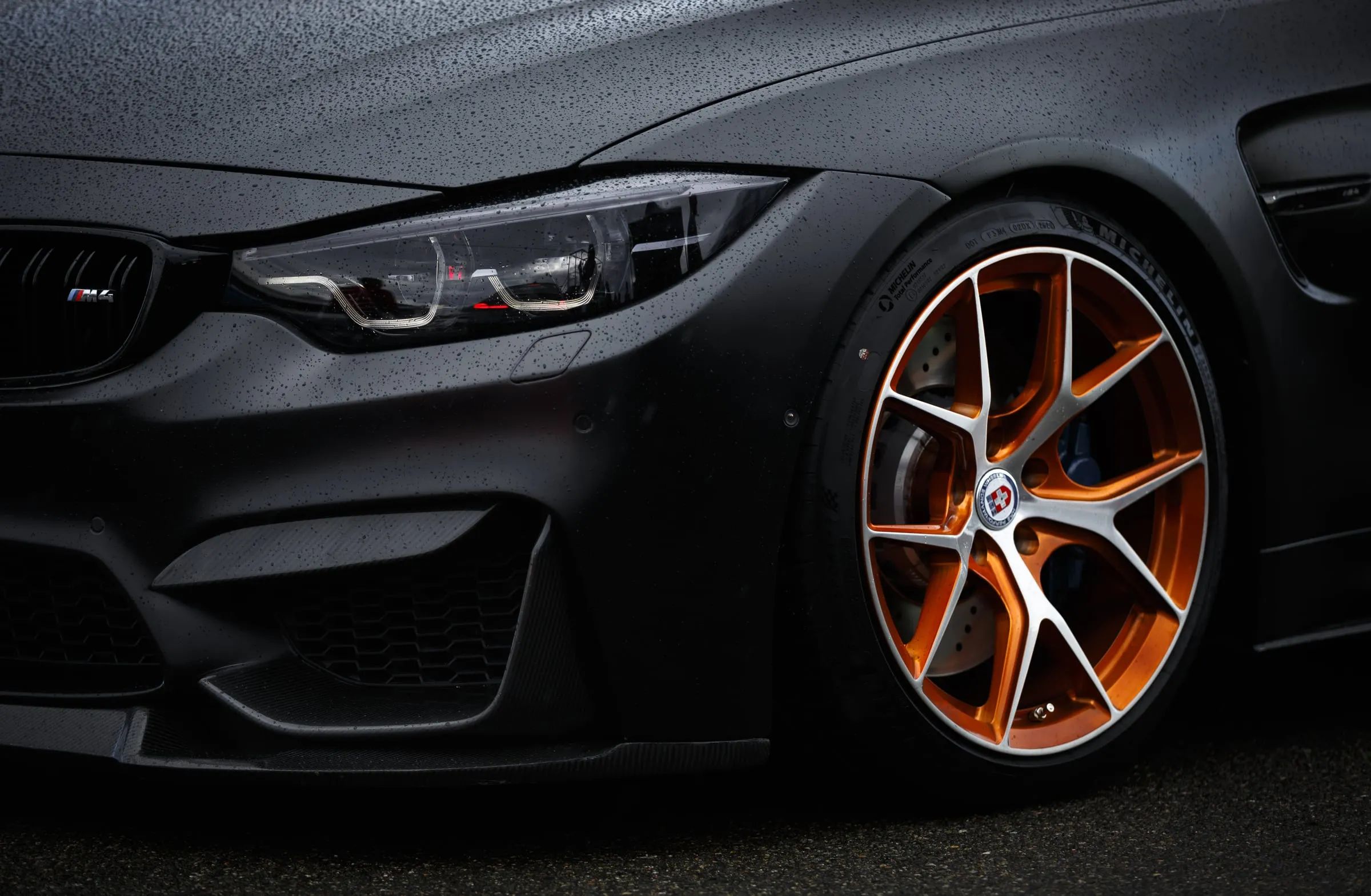 The front corner of a matte black BMW M4 on bronze wheels, wet from rain